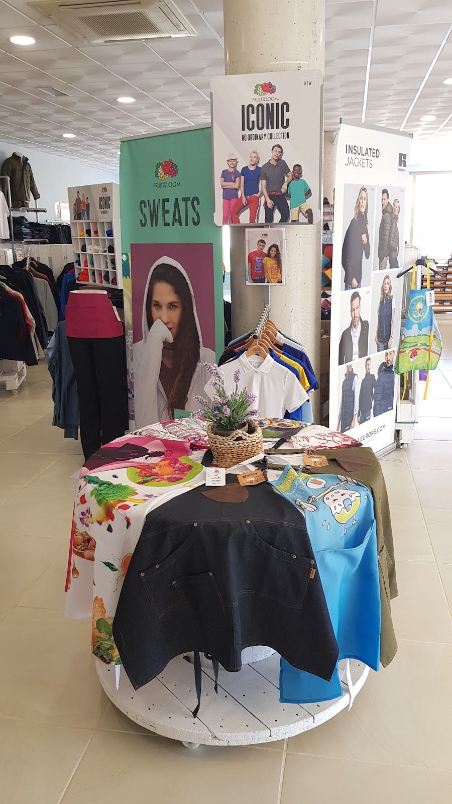 Mesa de exposición en tienda con delantales de cocina vaqueros y estampados junto a polos de trabajo
